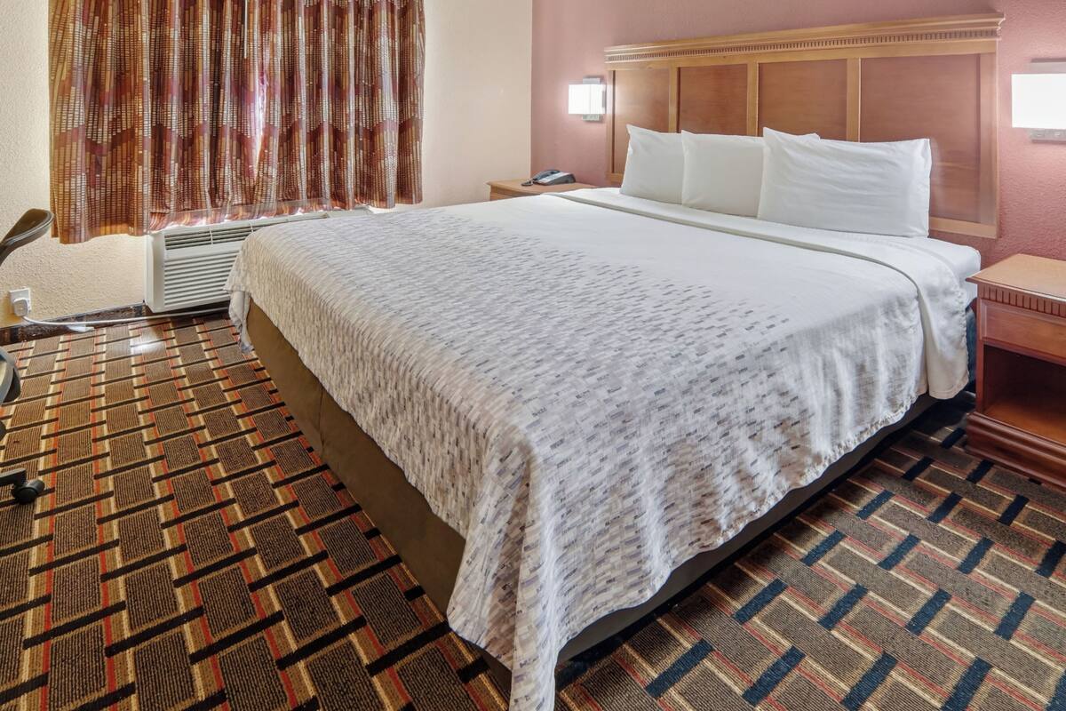 A hotel room with a large bed, a bedside table, a chair, an air conditioning unit, and patterned carpet.