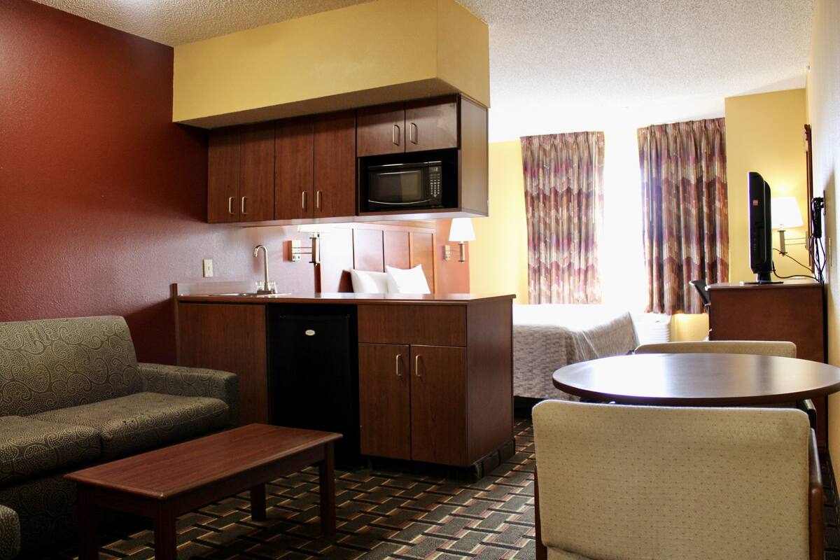A compact hotel room with a kitchenette, a sofa, a dining table, and a single bed.