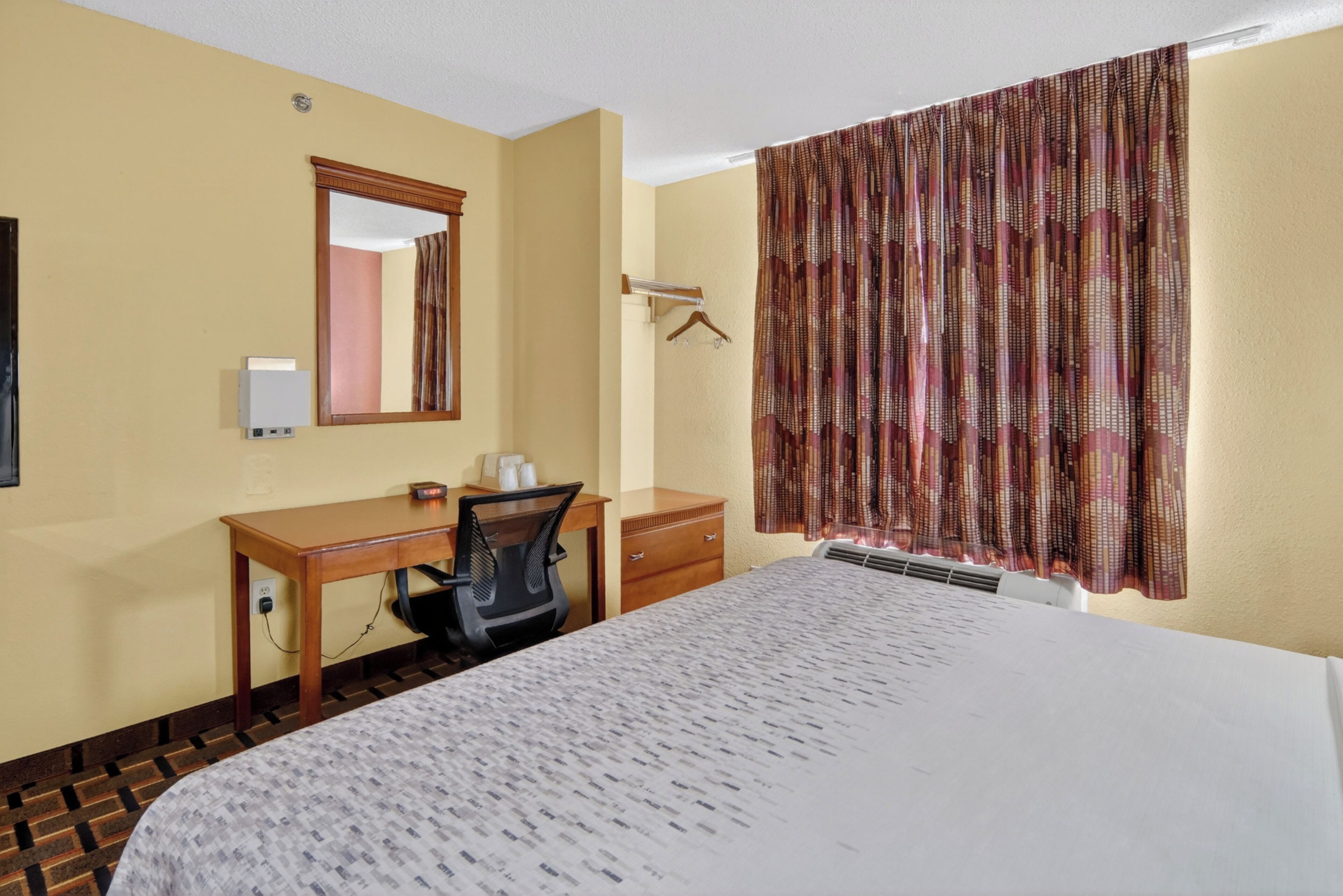 Een hotelkamer met een bed, bureau, stoel, linnenkast, spiegel en een raam met gordijnen.