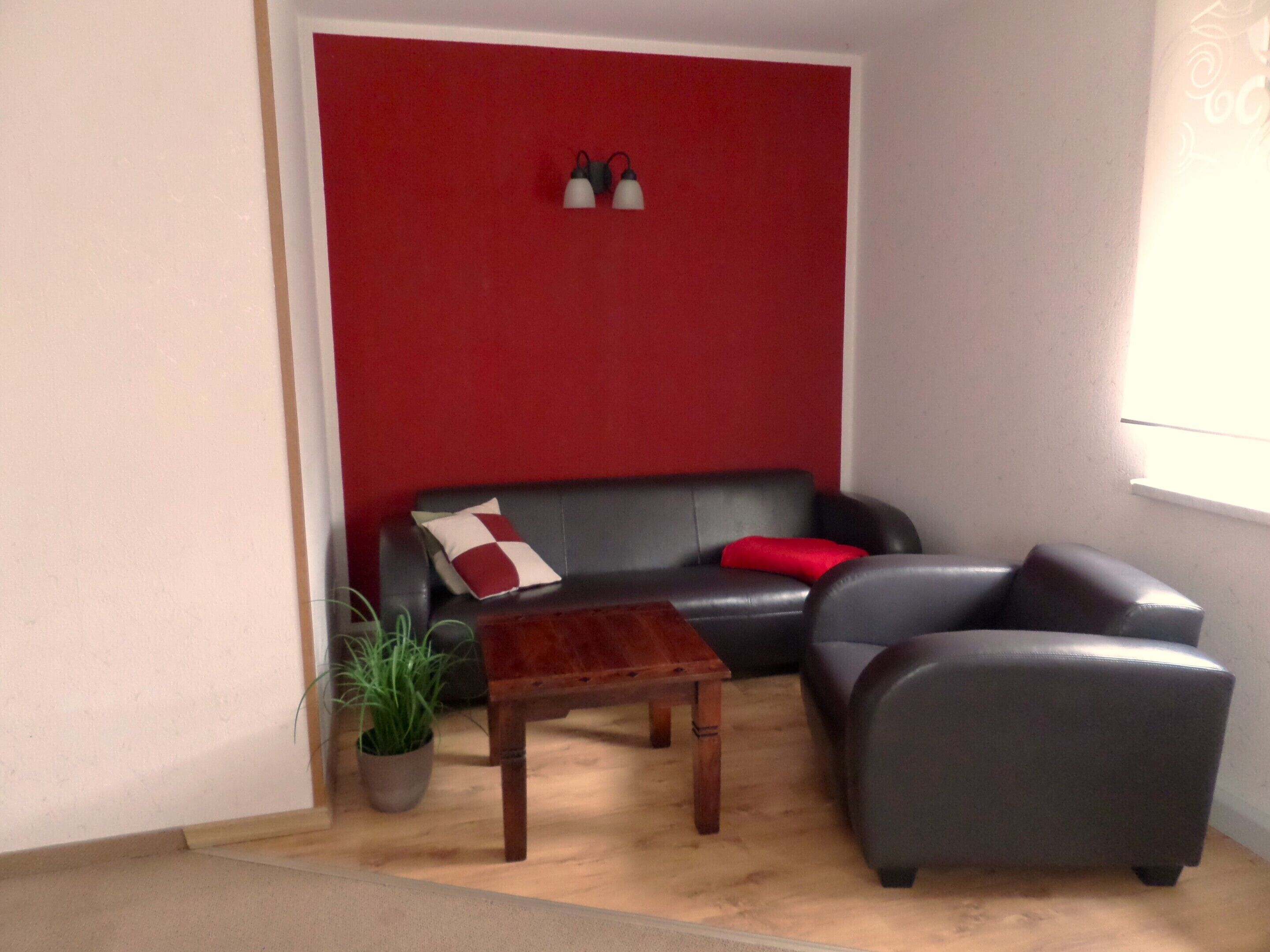 Uma sala de estar com parede de destaque vermelha, um sofá de couro preto, uma poltrona cinza, uma mesa de centro de madeira e um vaso com planta.
