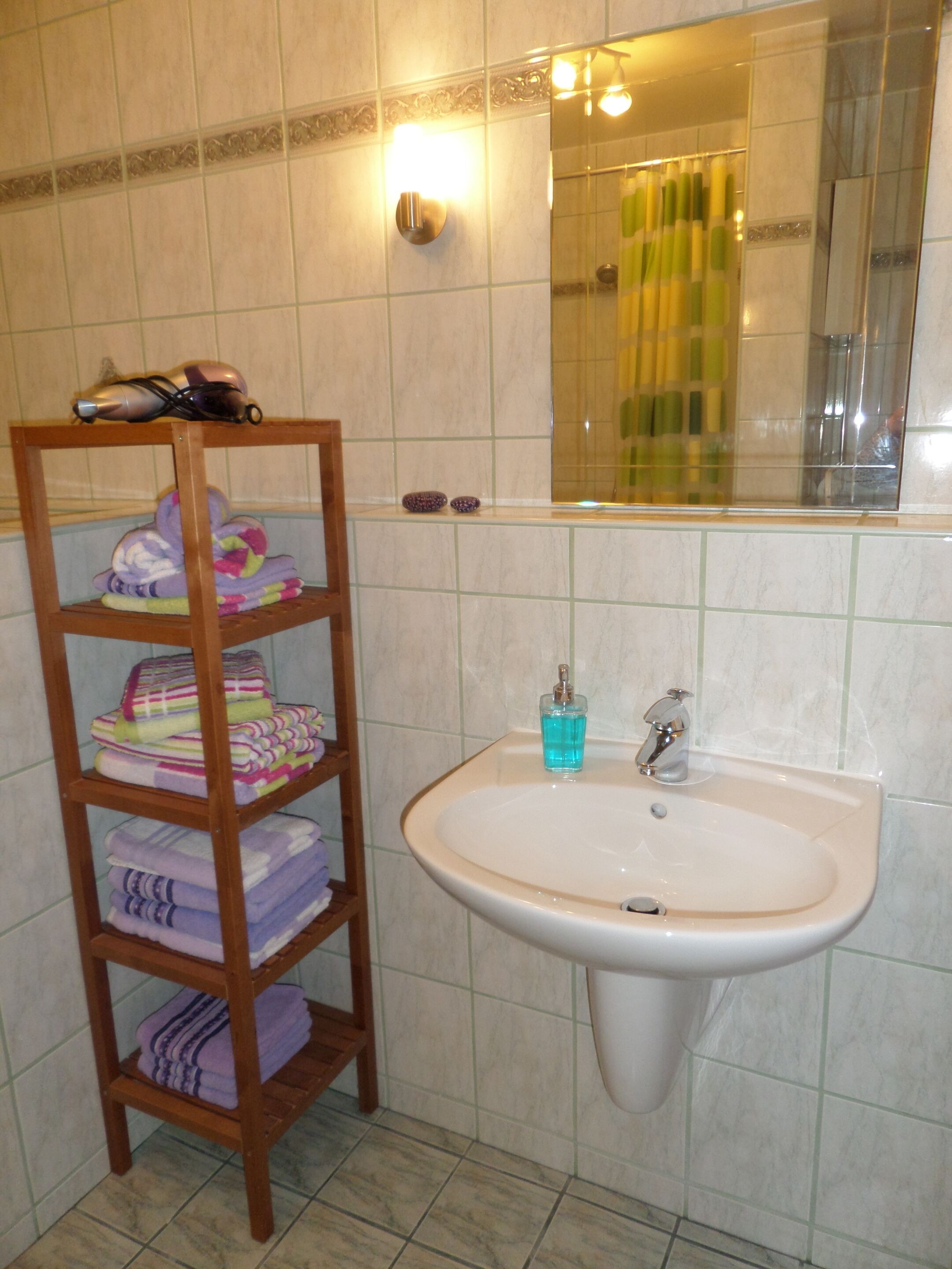A bathroom with a wooden shelf holding towels, a sink with a blue soap dispenser, and a mirror reflecting a shower curtain.