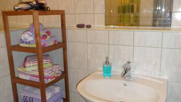 A bathroom with a wooden shelf holding towels, a sink with a blue soap dispenser, and a mirror reflecting a shower curtain.