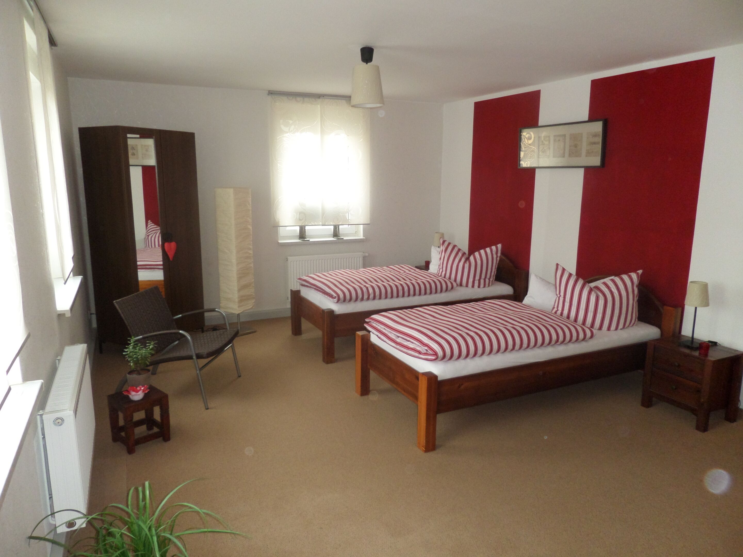 A bedroom with two single beds, a chair, a small table, a radiator, a window with curtains, and a red and white wall design.