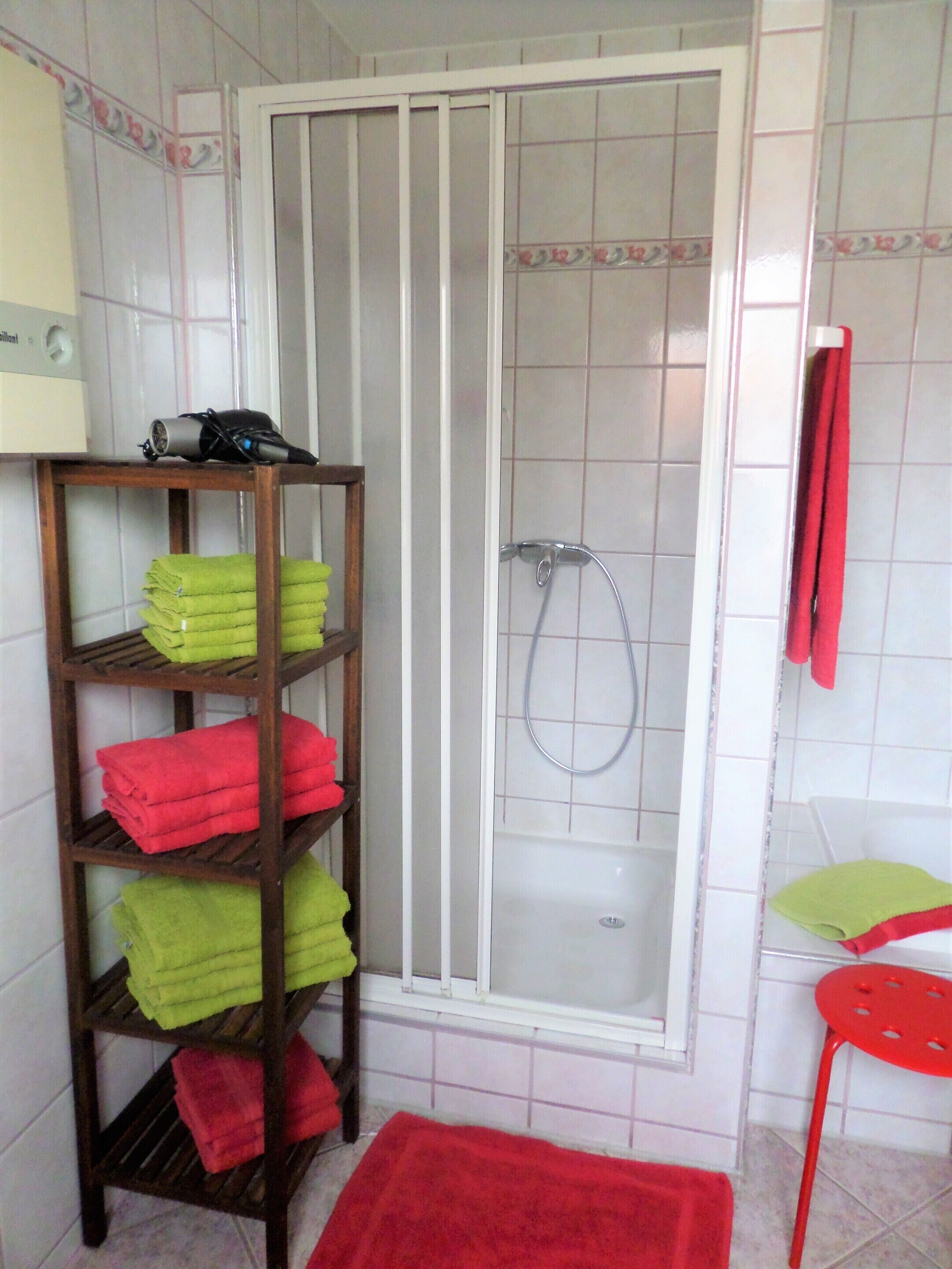 A bathroom with a shower, a wooden shelf with towels, and a red chair.