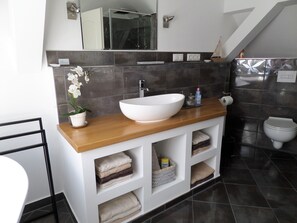 A modern bathroom with a white sink, wooden countertop, and a large mirror above. There are shelves with folded towels and a small potted plant.