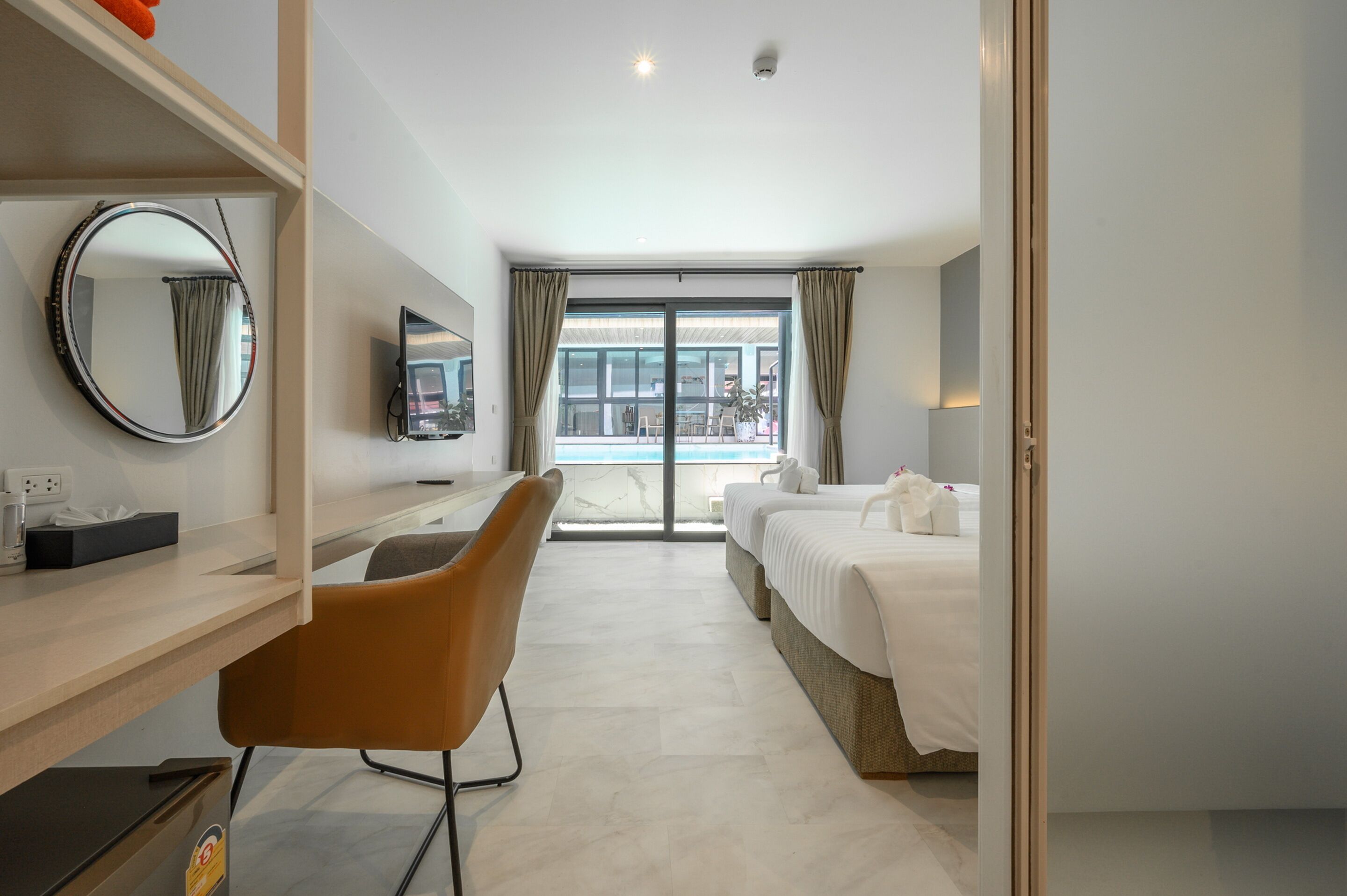 A hotel room with a desk, chair, two beds, a round mirror, and a TV.
