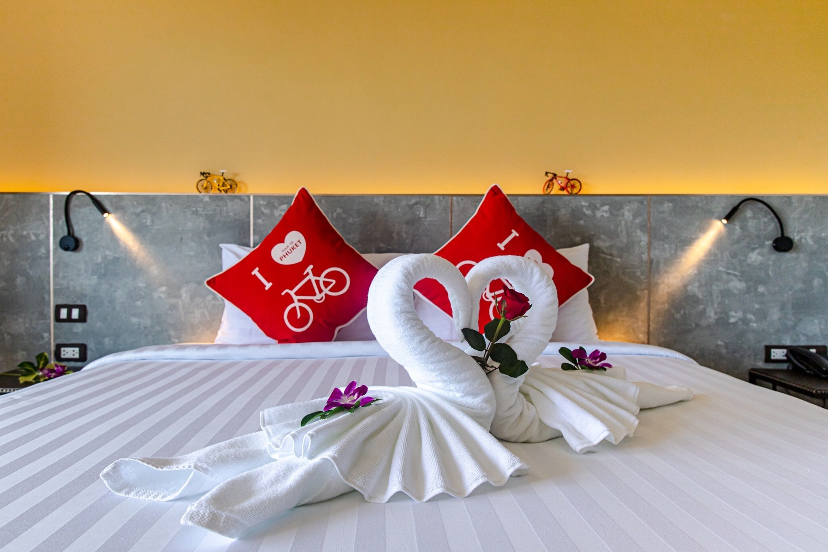Una cama bien hecha con una colcha a rayas, dos almohadas rojas con diseños de bicicletas y rosas, y un par de toallas blancas dispuestas en forma de cisnes con una flor morada.