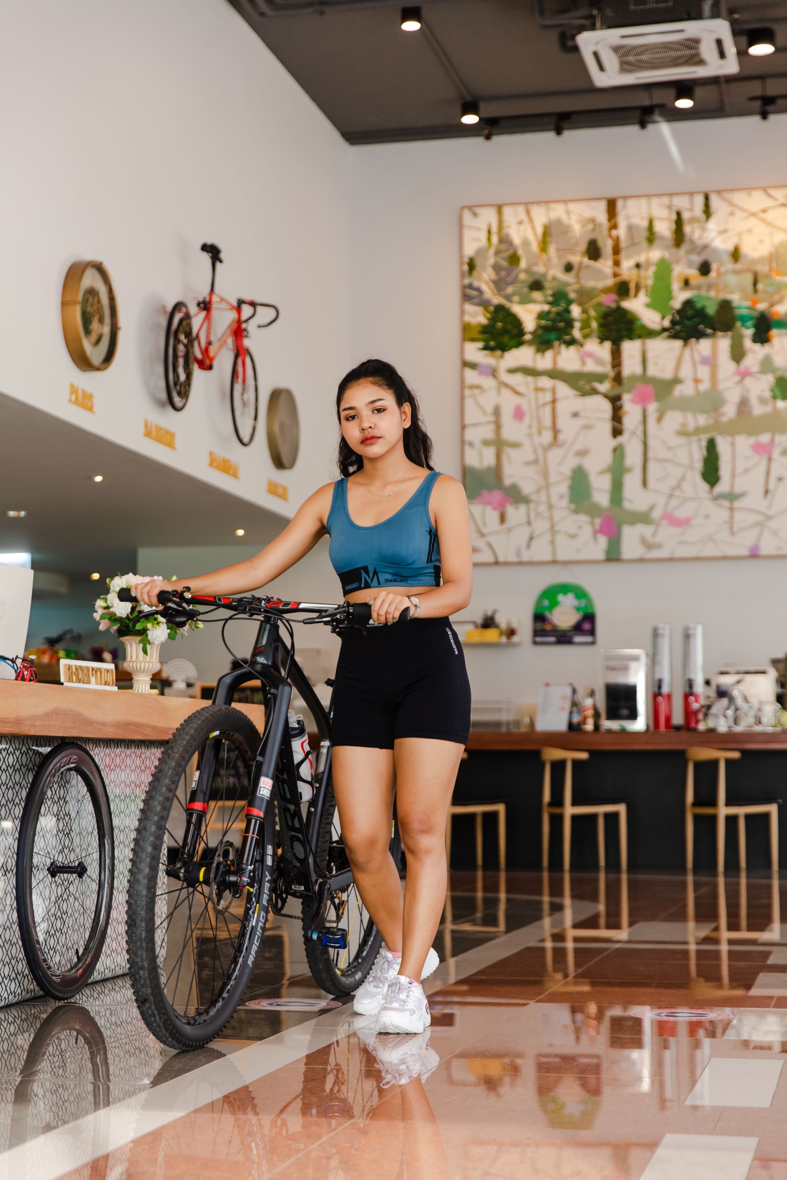 Une personne se tient à côté d’un vélo dans un espace intérieur moderne, doté d’un sol poli, d’un comptoir en bois et d’un mur orné d’objets décoratifs et d’un grand tableau abstrait.