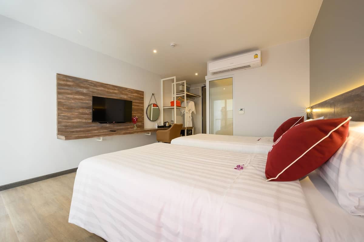 A hotel room with two beds, a TV mounted on a wooden panel, a wardrobe, a mirror, and an air conditioning unit.