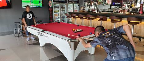 Een biljarttafel met een rode tafeldoek, een poolkeu en ballen. Er staan krukken bij en in de achtergrond is een bar te zien met flessen en glazen.