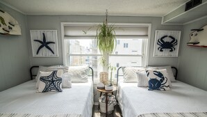 Habitación con dos camas, decoración de arte mural con estrellas de mar y cangrejos, una planta colgante y vistas a edificios a través de amplias ventanas.