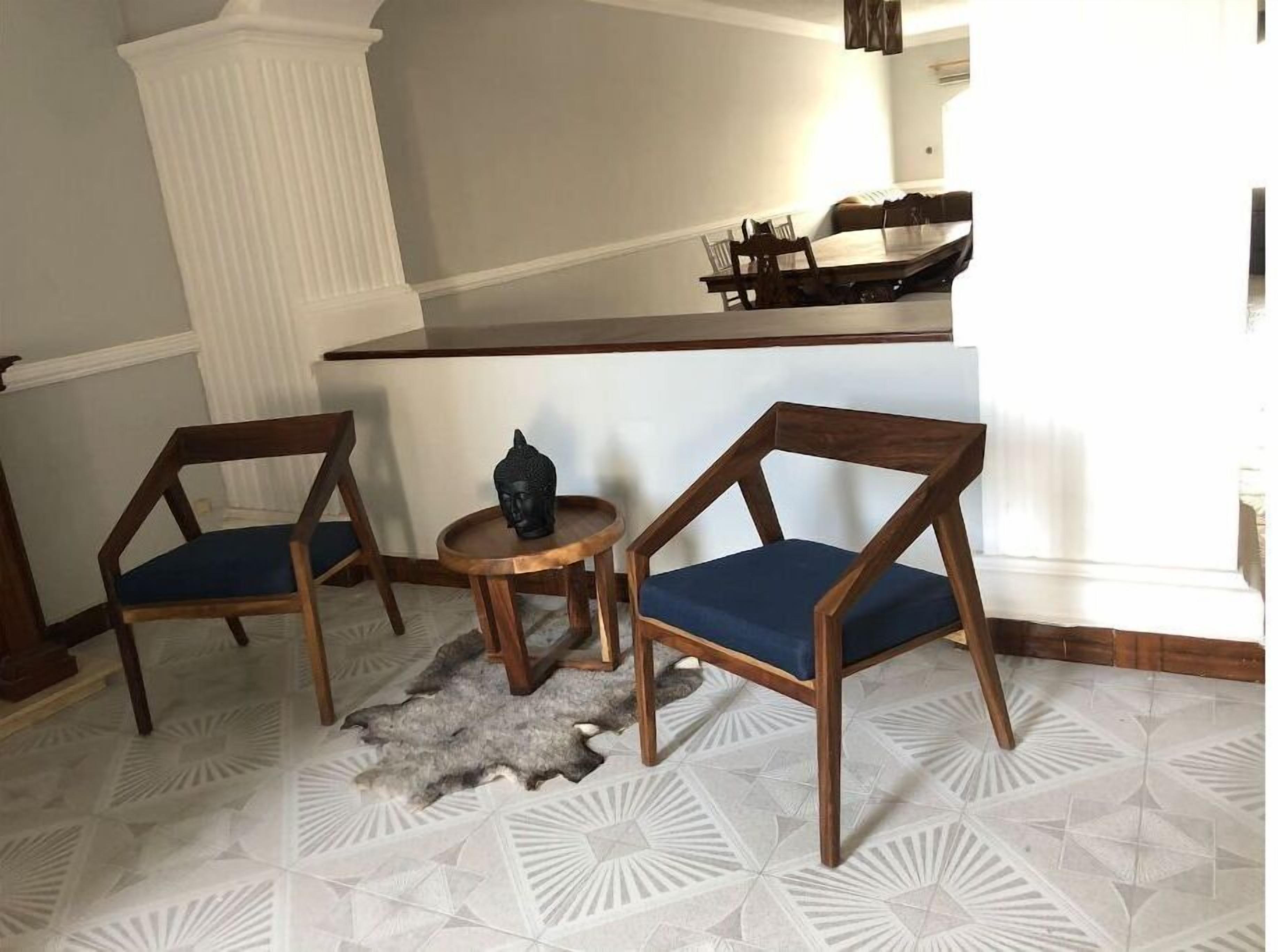 A room with two wooden chairs with blue cushions, a small round table with a Buddha statue, and a fur rug on the patterned floor.