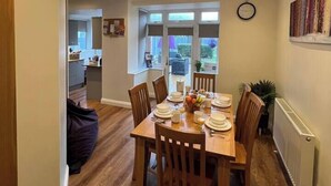 Uma área de refeições com mesa de madeira posta para quatro pessoas, rodeada por cadeiras de madeira. Há uma janela com vista para o exterior, um relógio na parede e um radiador à direita.