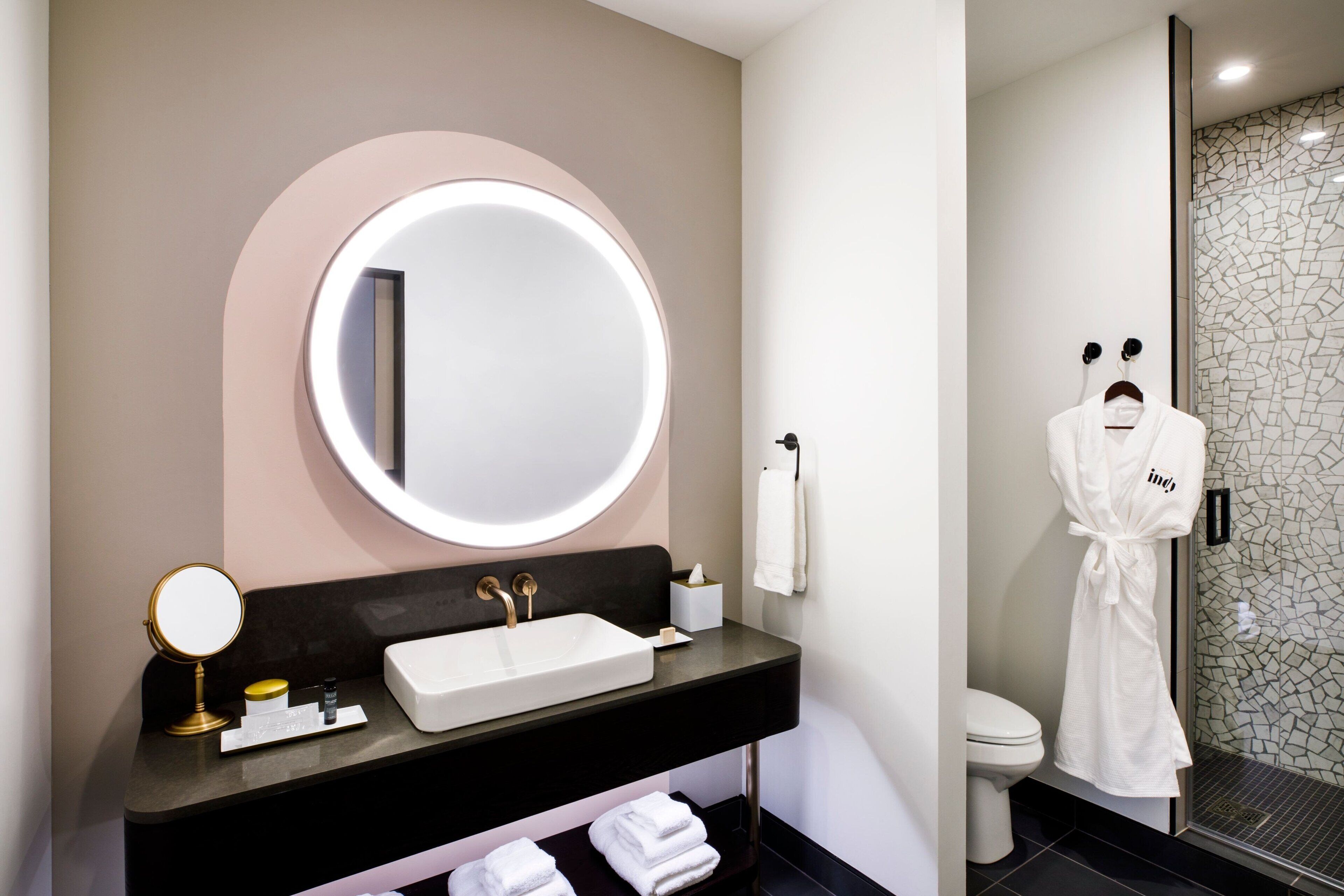 A modern bathroom with a round mirror, a rectangular sink, and a toilet. There is a robe hanging on a hook and a towel rack with folded towels.