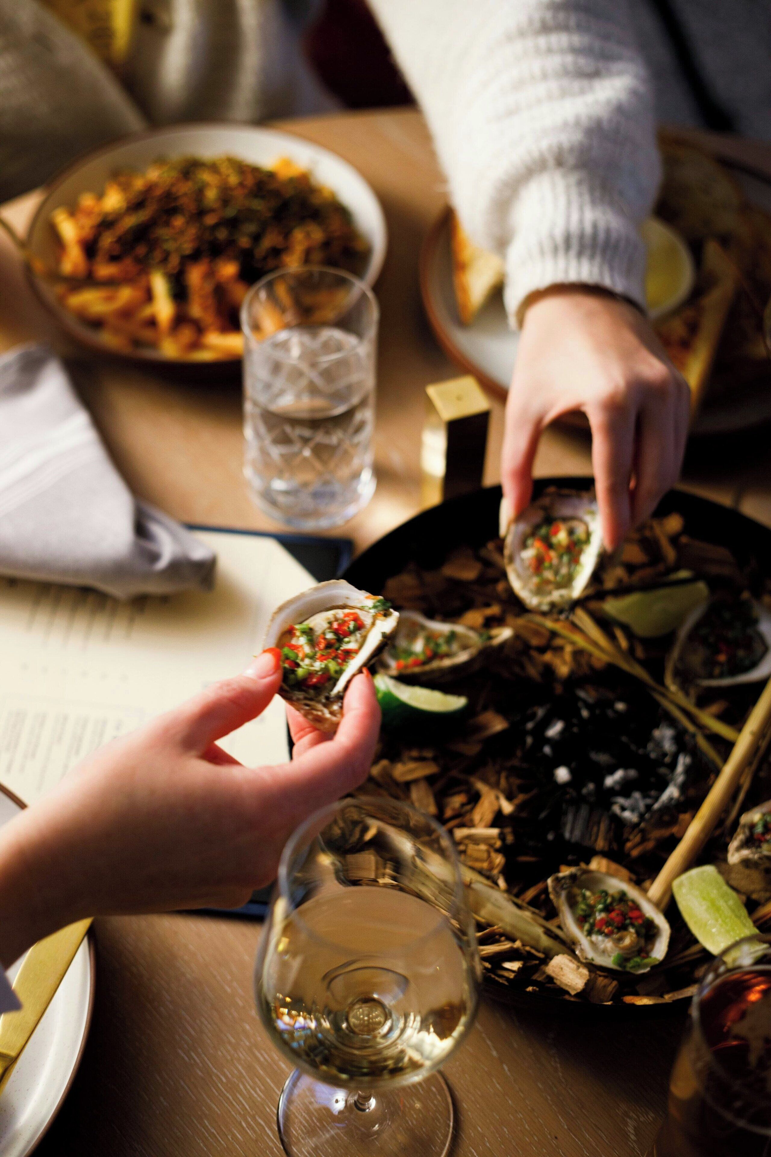 A dining table with a plate of food, a glass of water, and a person holding an oyster.