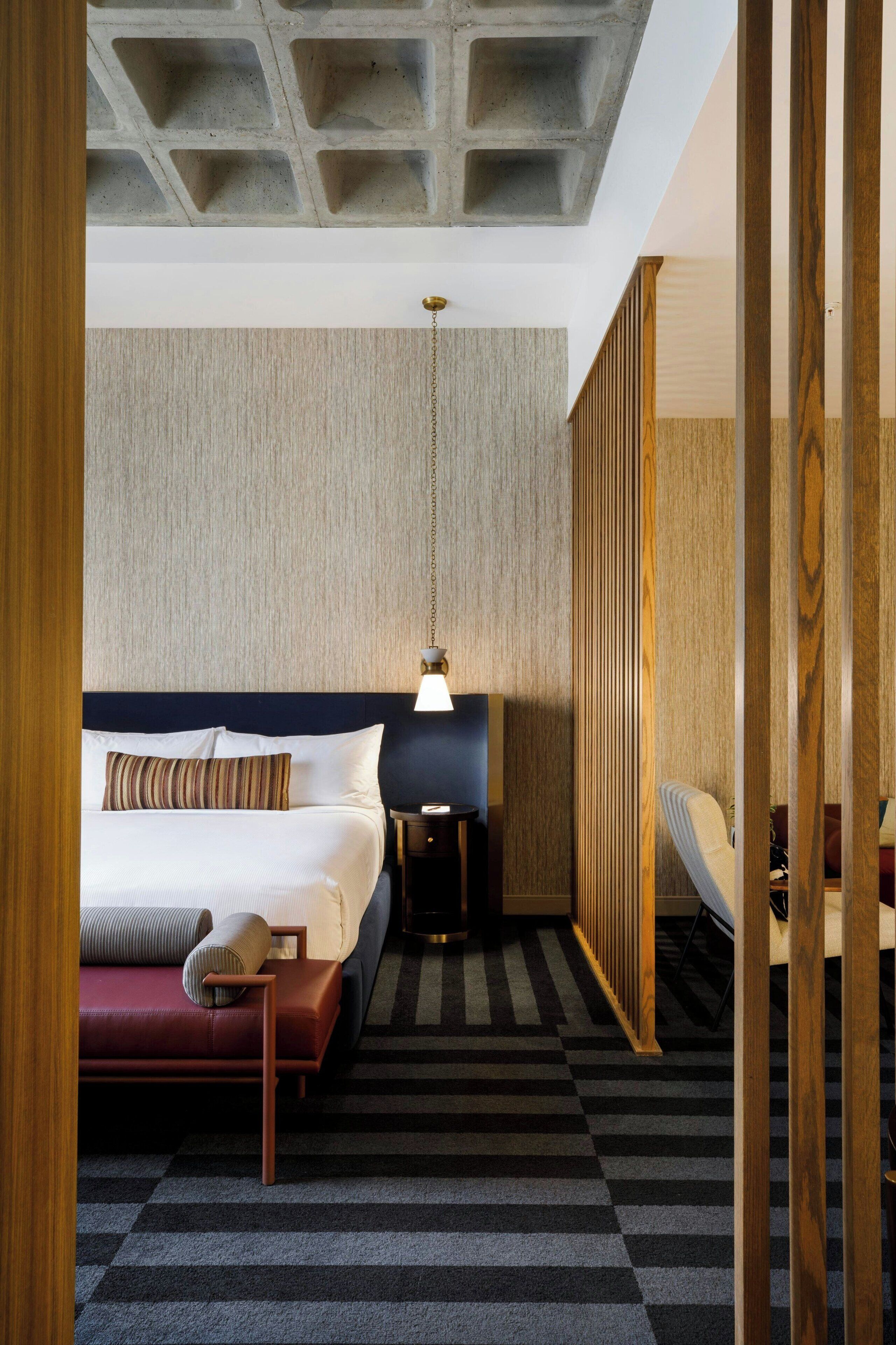 A hotel room with a large bed, a bedside table, a lamp, and a view of the outside through a wooden partition.