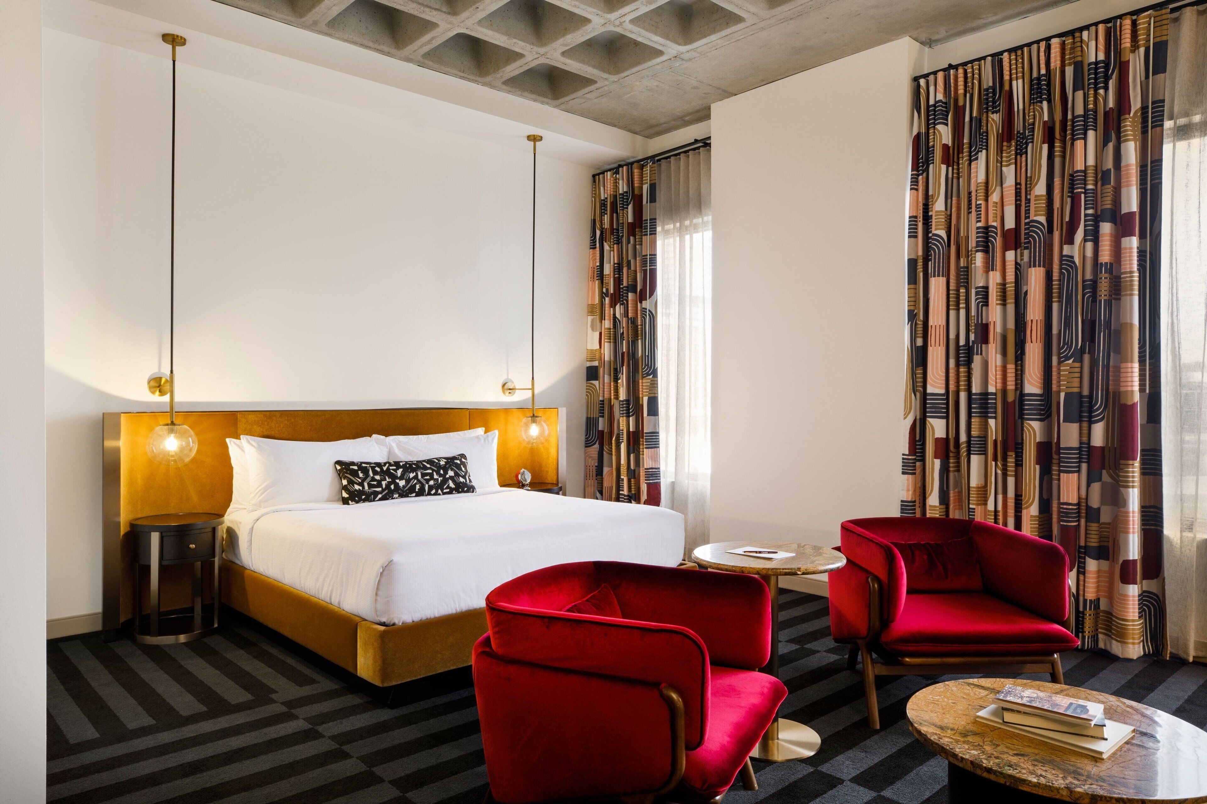 A hotel room with a large bed, two red armchairs, a small round table, and a patterned curtain.
