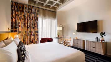 A hotel room with a large bed, a TV, a desk with a lamp, a red chair, and a patterned curtain.