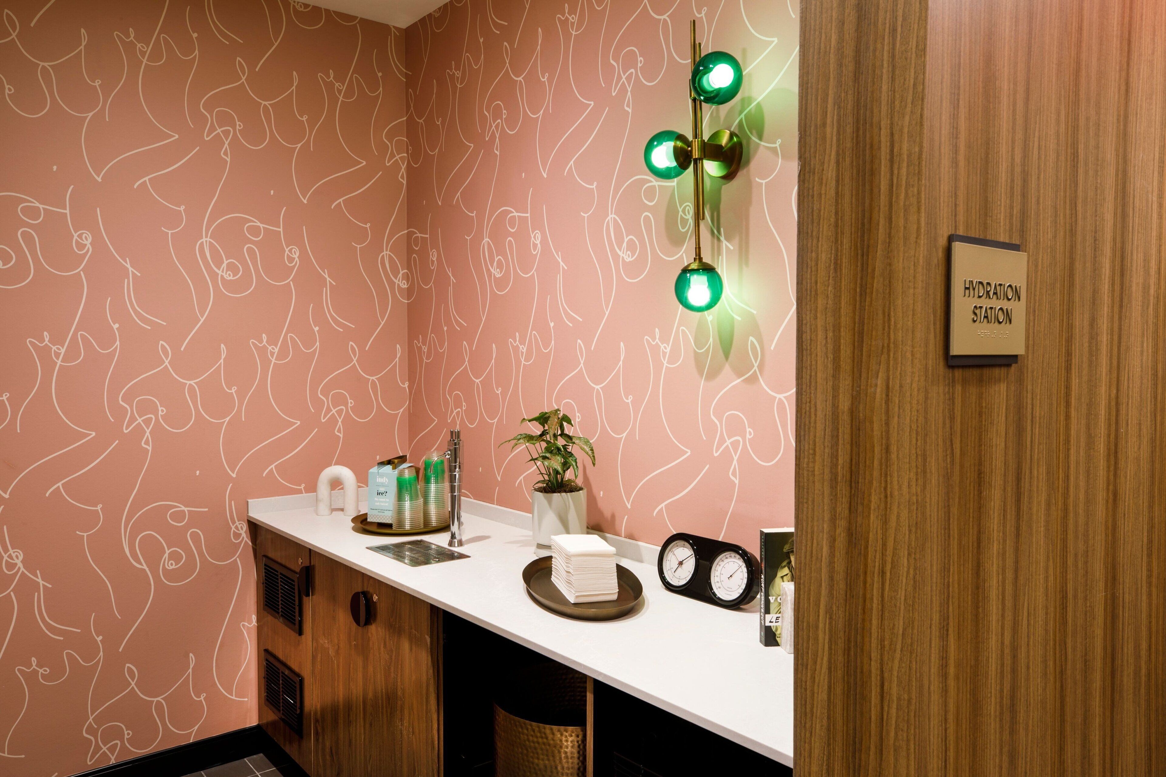 A bathroom with a pink patterned wall, a white countertop, a sink, a mirror, a clock, and a sign reading "HYDRATION STATION".