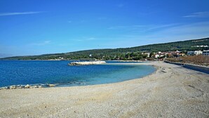 Uma praia de pedras com águas azul-claras, um litoral rochoso e colinas e edifícios ao fundo.