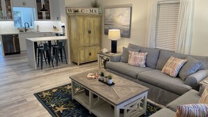 A modern living room with a grey sofa, a coffee table, and a kitchen area with white cabinets and a 'WELCOME' sign. - Swim Up Bar - Outdoor Paradise! (Delray Beach)