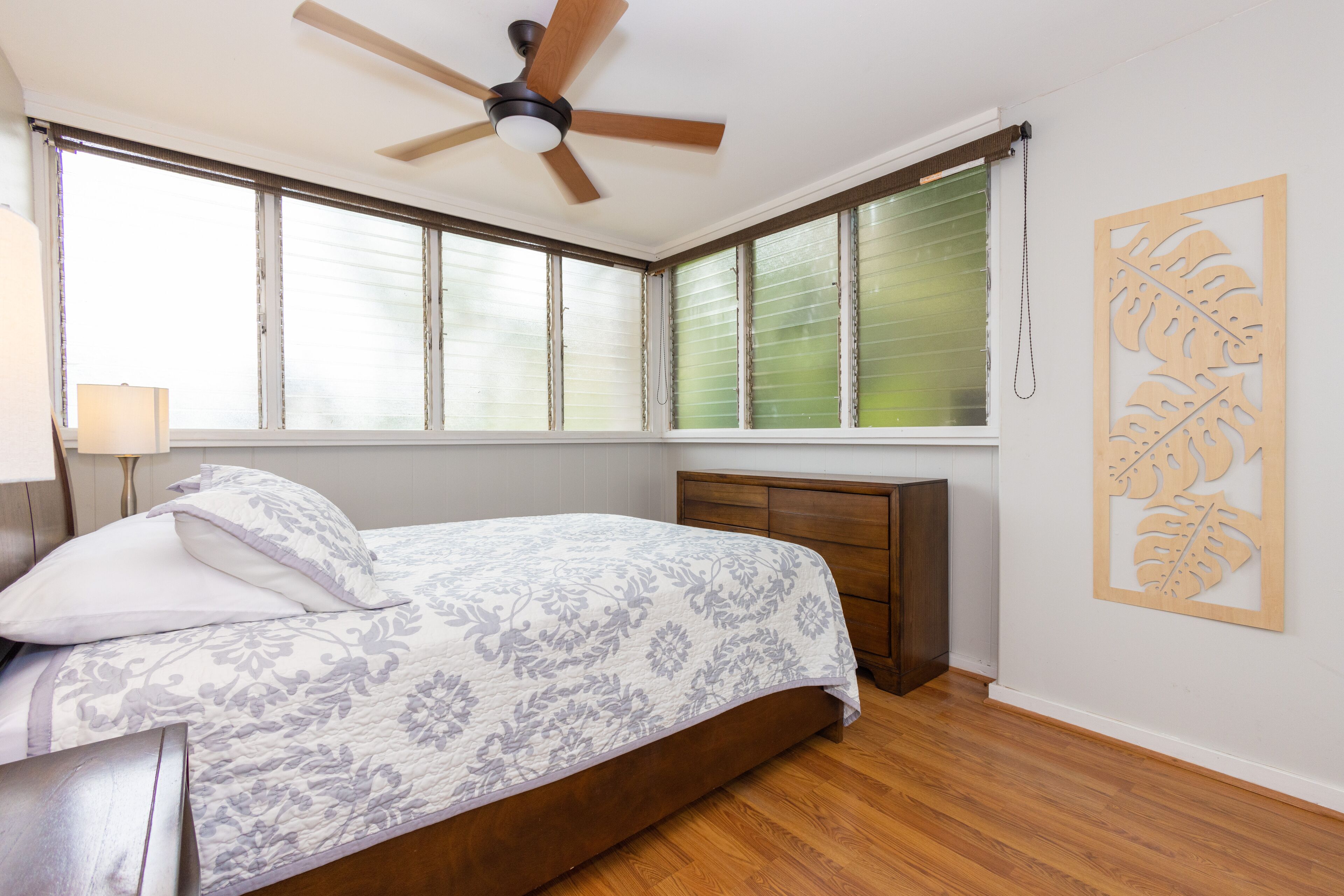 Ein Schlafzimmer mit einem Bett, einem Deckenventilator, einem Fenster mit Jalousien, einem Holz-Kopfende und einer Wanddekoration.