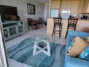 A living room with a glass coffee table, blue sofa, and a kitchen area with wooden cabinets and a refrigerator.