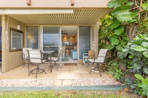 A covered patio with a dining area, sliding glass doors leading to a kitchen, and a lush green wall.