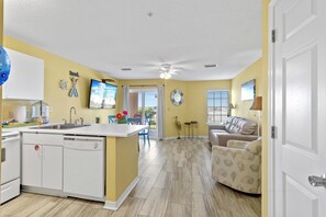 A compact kitchen with a dishwasher, a dining area with a table and chairs, a sofa, and a view through a glass door to the outside.