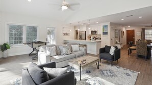 Ein geräumiges Wohnzimmer mit einer Couch, Sesseln und einem Couchtisch. Es gibt eine Küchenzeile mit modernen Geräten sowie einen Essbereich mit Tisch und Stühlen.