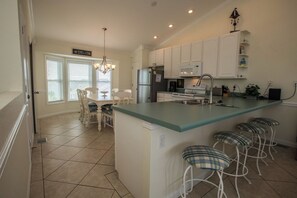 A kitchen with white cabinets, a central island with bar stools, a refrigerator, and a dining area with a table and chairs. - Sea Cliffs rental with elevator SUN-SUN Rental (Port Saint Joe)
