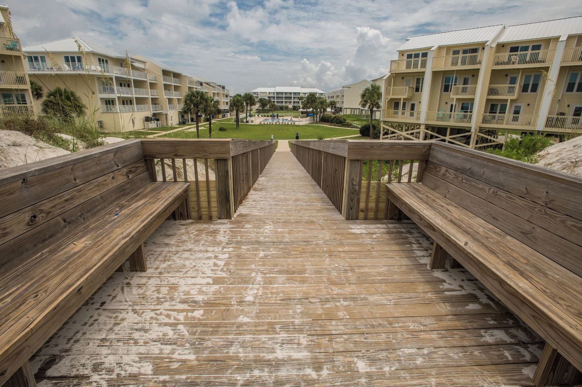 A wooden boardwalk leading to a beachfront hotel with multiple balconies and a pool area.