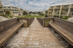 A wooden boardwalk leading to a beachfront hotel with multiple balconies and a pool area.