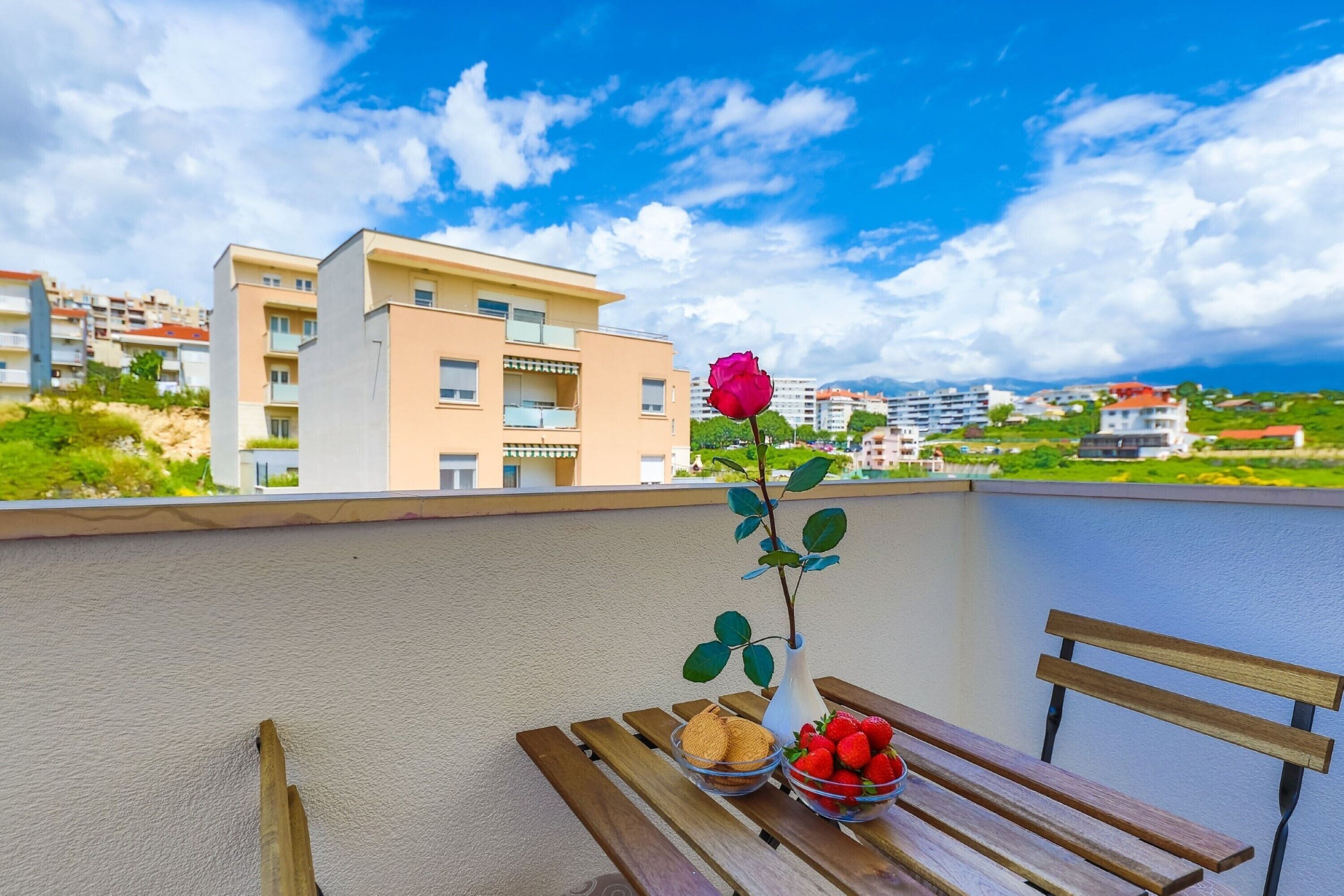 A balcony with a wooden bench, a vase with a single rose, and bowls of strawberries and cookies.