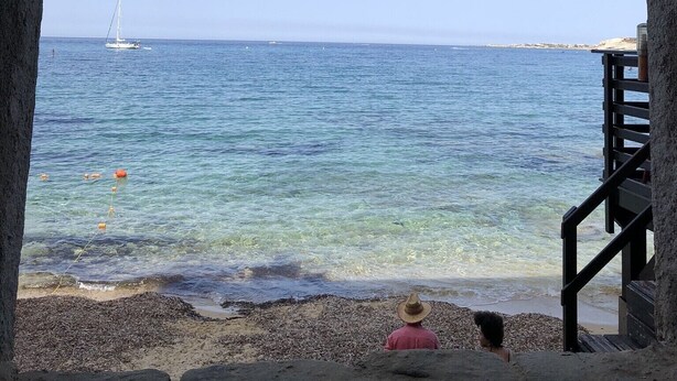 A stone archway overlooking a beach with clear water, a sailboat in the distance, and a person in a pink shirt and straw hat. - Apartment Benvenuti Calvi the beach with feet with 3 min, terrace, carpark, swimming pool (Calvi)