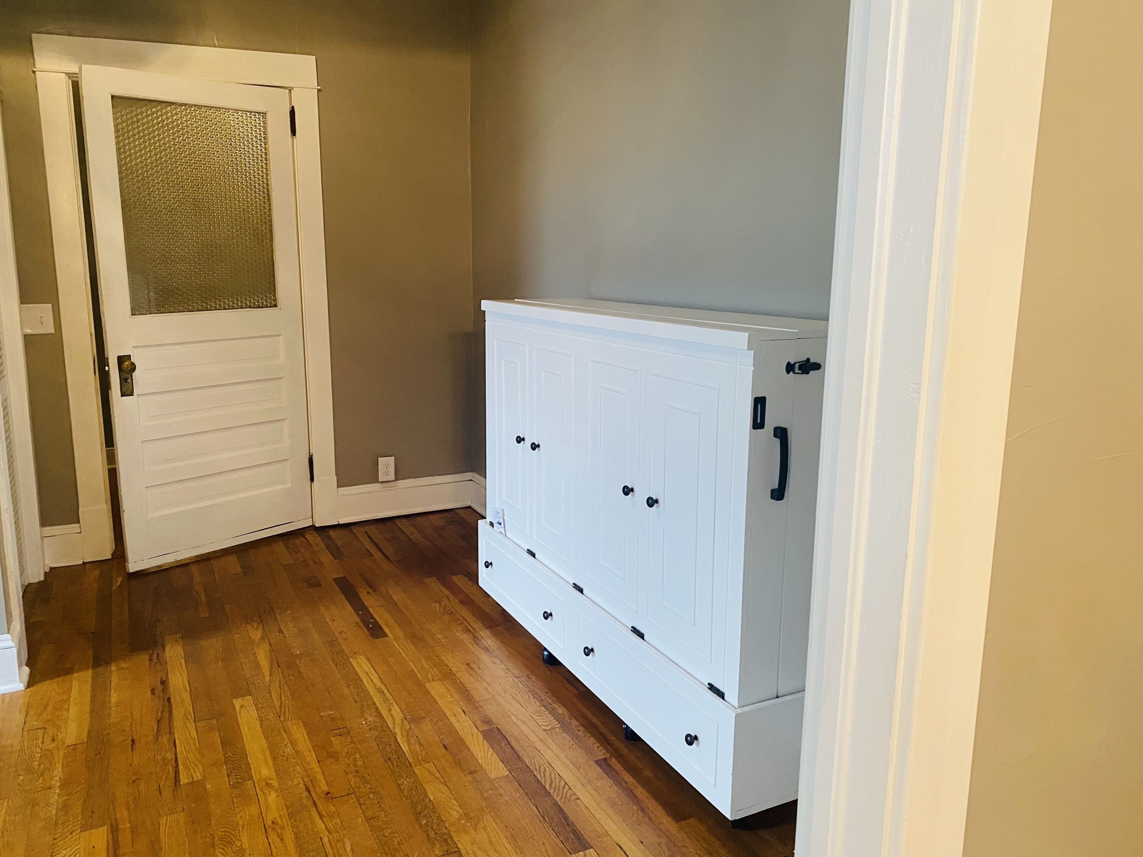 A room with wooden flooring, a white cabinet with drawers, and a door with a frosted glass panel.