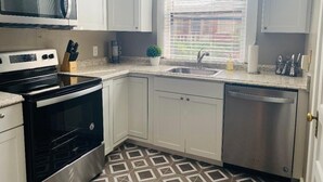 A compact kitchen with white cabinets, a microwave, a stove, a dishwasher, and a window with blinds.
