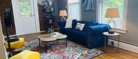 Una sala de estar con un sofá azul, un pouf amarillo, una mesa de centro y una alfombra.
