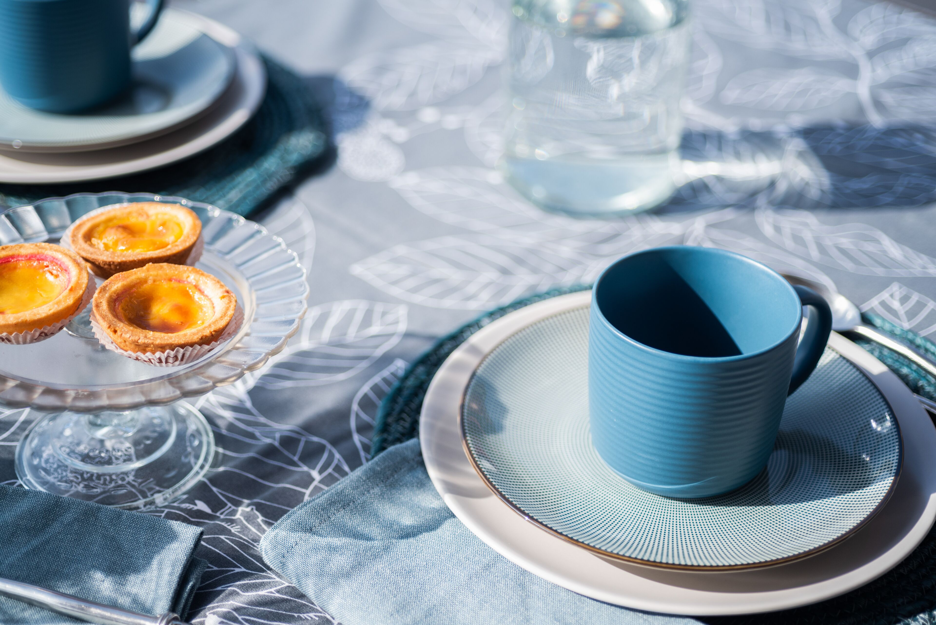 Una mesa con una plancha de vidrio que sostiene tres pequeños pasteles, una taza azul sobre un plato y un vaso transparente.