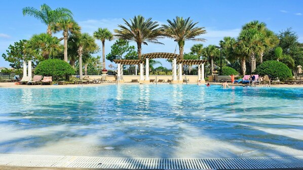 Relax by the pool surrounded by lush greenery and palm trees, with lounge chairs and umbrellas providing shade under a pergola.