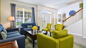 Una sala de estar con un sofá azul, sillones verdes, una mesa de centro con flores amarillas y una escalera con barandillas blancas.