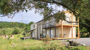 Uma casa de madeira de dois andares com varanda, cercada por vegetação e um caminho de cascalho.