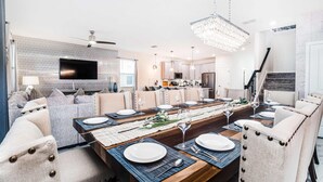 A modern dining room with a long wooden table set for a meal, a television in the background, and a chandelier above. - Avatar Game Room + Harry Potter BR w/ Pool~Spa (Kissimmee)