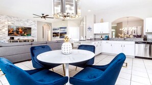 A modern living room with a grey sofa, a round table with blue chairs, a TV mounted on a white brick wall, and a kitchen with white cabinets and stainless steel appliances. - Close2Parks, 7BR, Themed room @ Windsor Hill! (Kissimmee)