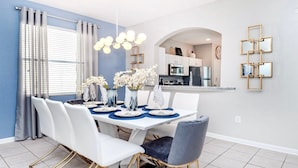 A dining area with a white table set for six, blue placemats, white chairs, and a vase of white flowers. - Close2Parks, 7BR, Themed room @ Windsor Hill! (Kissimmee)