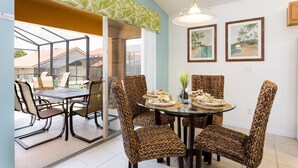 A dining area with a glass table surrounded by woven chairs, a hanging light fixture, and framed pictures on the wall.
