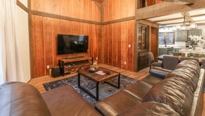 Un salón espacioso con una gran pared de madera, un televisor de pantalla plana, una mesa de centro, sofás de cuero y una zona de comedor al fondo.