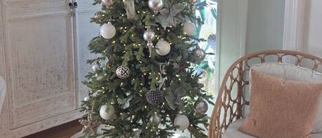 Un árbol de Navidad con adornos y luces, una silla blanca con cojín, y una alfombra redonda de color blanco.