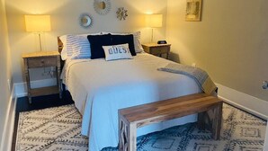 A neatly arranged bedroom with a bed, two bedside tables, a nightstand, a bench, patterned rugs, and wall decor. - Amazing 1 Bedroom Apartment, Downtown Jonesborough (Jonesborough)