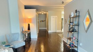 A hallway with wooden flooring, a grey armchair, a small table with a lamp, and a metal shelf with various items. - Amazing 1 Bedroom Apartment, Downtown Jonesborough (Jonesborough)